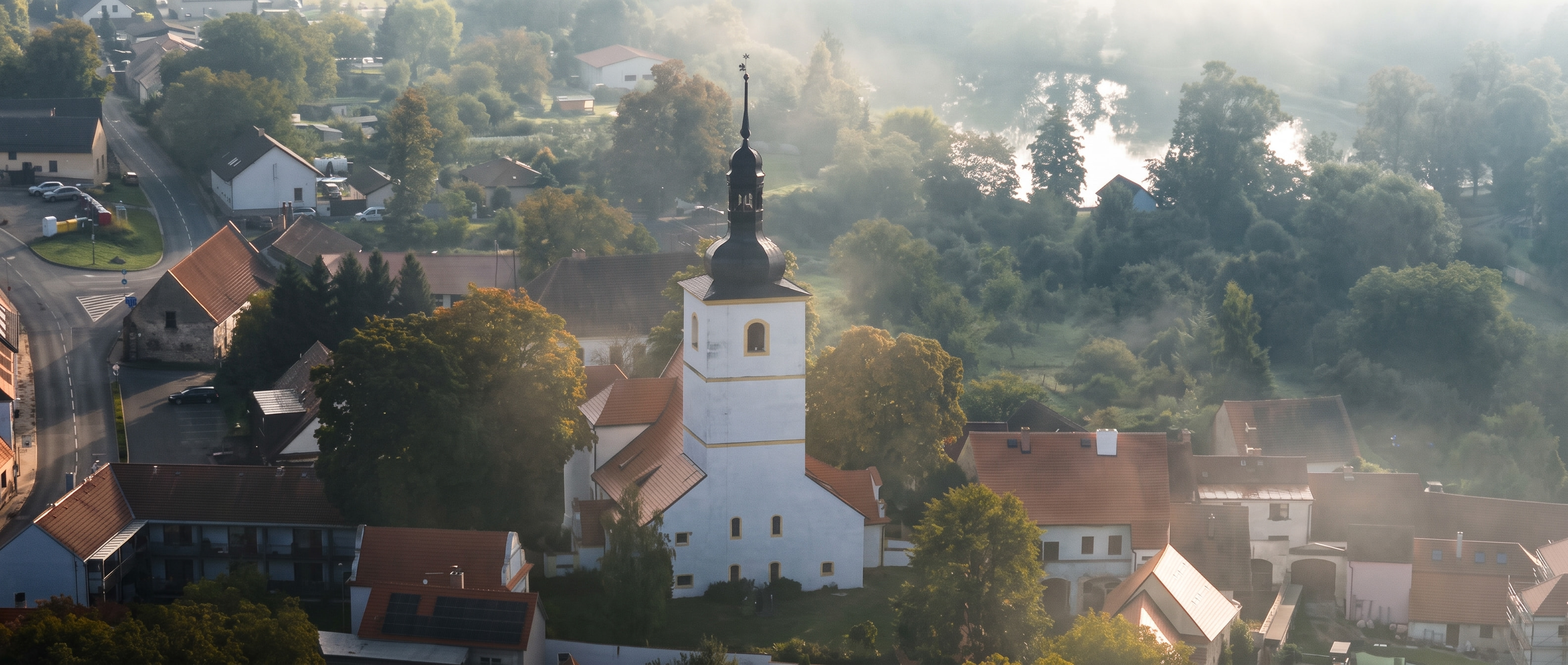 Radomyšl brzy ráno: bílá věž kostela nad střechami domů, mlha nad stromy